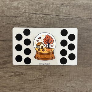 May include: A white rectangular sticker with a fall-themed snow globe design. The snow globe features a house, tree, and pumpkin. The sticker has black circles on each side and the text "Spooky Budgets" at the bottom.