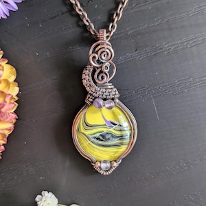 May include: A handcrafted copper pendant necklace featuring a vibrant, abstract design in yellow, black, and purple. The pendant is wire-wrapped with intricate detailing and includes small purple beads. The necklace has a copper chain.
