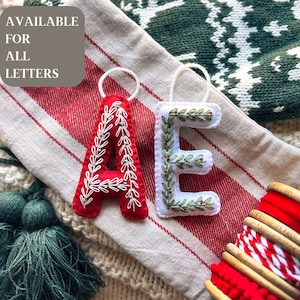 May include: Two felt Christmas ornaments, one red with white embroidery and the letter A, the other white with green embroidery and the letter E. The ornaments are on a striped fabric background. Available for all letters.
