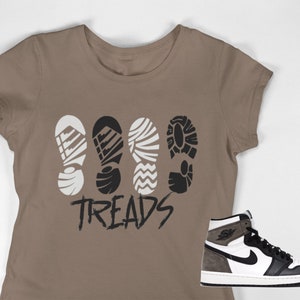 Puede incluir: Una camiseta marrón con un gráfico de cuatro patrones de suela de zapato y la palabra "TREADS" en negro. Una zapatilla de deporte de caña alta blanca y negra se muestra debajo de la camiseta.