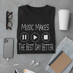 Pode incluir: Camiseta preta com o texto "MUSIC MAKES THE BEST DAY BETTER" e símbolos musicais. A imagem também inclui óculos, um smartphone, um par de tênis, um copo de café e um jornal.
