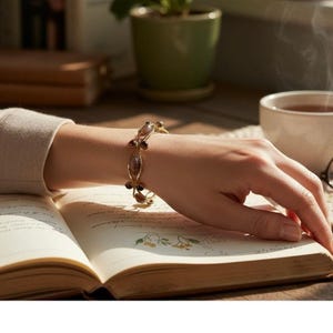 May include: A gold-toned bracelet with oval and round gemstones is displayed on a wrist. The bracelet features a delicate design with alternating light brown and dark brown stones. The wrist rests on an open book with floral illustrations, suggesting a theme of elegance and vintage style.