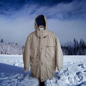 May include: A tan winter parka with a hood and multiple pockets. The jacket has a dark blue lining in the hood and a button and zipper closure. The background shows a snowy landscape with trees and a cloudy sky.