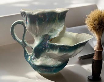 May include: A ceramic shaving scuttle with a handle and a built-in bowl, featuring a pearlescent finish with shades of blue and white. A shaving brush with a wooden handle is next to the scuttle.