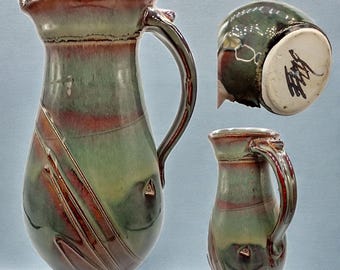 May include: A handmade ceramic pitcher with a large handle and a flared rim. The pitcher features a blend of green, red, and brown glazes with diagonal lines. A smaller matching pitcher is also shown. The bottom of the pitcher has a signature.