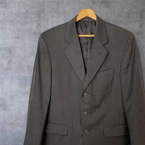 Könnte beinhalten: Ein dunkelgrauer Nadelstreifenblazer mit einem einzigen Knopfverschluss und zwei Taschen. Der Blazer hängt an einem Holzbügel.
