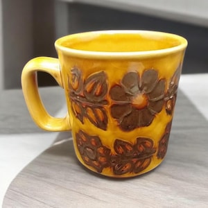 May include: A yellow ceramic mug with a brown floral design. The mug has a rounded handle and a wide rim.