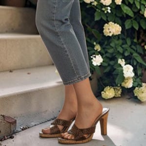 May include: A pair of brown snakeskin-patterned mules with a cork-style platform and high heel, paired with cropped grey denim jeans. The footwear features a wide, crisscross strap design across the top of the foot, providing an open-toe style.