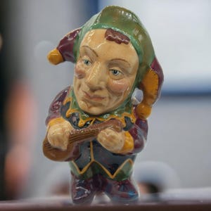 Puede incluir: Figura de cerámica de un bufón, con un rostro detallado y un disfraz colorido. El bufón sostiene un pequeño instrumento de cuerda, con un sombrero verde, morado y amarillo. La figura tiene un aspecto vintage y artesanal.