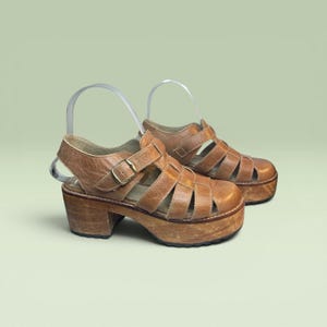 May include: A pair of tan leather platform sandals featuring a caged upper design with multiple straps, a rounded toe, and an adjustable ankle strap with a gold-toned metal buckle. The shoes have a thick, chunky wooden block heel and platform sole.