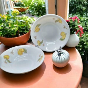 Könnte beinhalten: Weiße Keramikschalen mit Zitronenmuster. Die Schalen sind mit gelben Zitronen und grünen Blättern verziert. Ein kleiner, weißer Keramik-Kürbis mit schwarzen Linien ist ebenfalls vorhanden. Die Schalen stehen auf einem runden, orangefarbenen Tisch.
