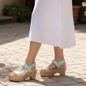 May include: A pair of platform sandals featuring a light blue ankle strap with a silver metal buckle, a white front strap, and a thick wooden sole. The sandals are worn with a white skirt, showcasing the light blue and white color combination.