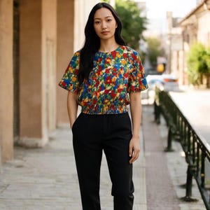 Könnte beinhalten: Eine Frau trägt ein Oberteil mit Blumenmuster, kurzen Ärmeln und eine schwarze Hose. Das Oberteil hat ein lebendiges Muster aus roten, gelben und blauen Blumen auf dunklem Hintergrund. Die Hose ist einfarbig schwarz.