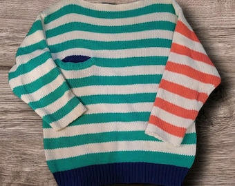 May include: A striped sweater with a white and green horizontal stripe pattern. One sleeve is a solid coral color, and the other sleeve has a white and coral stripe pattern. The sweater has a blue trim around the neck and bottom.