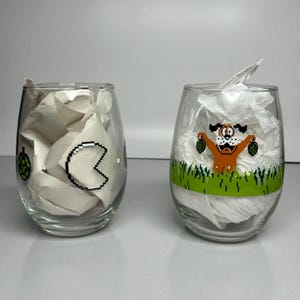 May include: Two clear stemless wine glasses with colorful pixelated designs. One glass features a Pac-Man ghost, and the other has a dog holding two ducks. Both glasses are filled with white tissue paper.