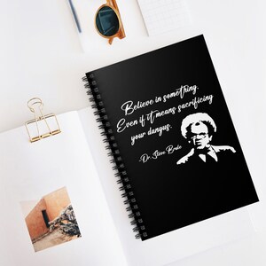 Könnte beinhalten: Schwarzes Spiralnotizbuch mit dem Zitat "Believe in something. Even if it means sacrificing your dangus." und einem weißen Porträt von Dr. Steve Brule. Das Notizbuch liegt offen auf einer weißen Oberfläche.