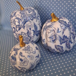 May include: Three decorative pumpkins featuring a white base with a blue floral pattern and metallic gold stems. The pumpkins vary in size and shape, with one tall, rounded pumpkin and two smaller, squatter pumpkins, all displaying intricate botanical designs.
