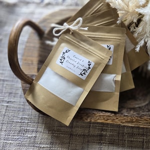 Puede incluir: Un montón de bolsas de papel marrón con una etiqueta blanca que dice "Curved & Chiseled Scented Laundry Soap" con un diseño floral. Las bolsas están atadas con cuerda y están sobre una bandeja de madera.