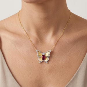 May include: A gold chain necklace featuring a butterfly pendant. The butterfly has yellow gold wings, a central pear-shaped red gemstone, and white metal edges encrusted with small clear stones that outline the entire wing structure.