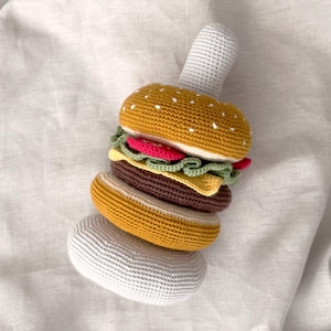 May include: A crocheted hamburger toy with a yellow bun dotted with white, a brown patty, and red, green, and yellow fillings. The toy includes a gray handle and a bottom bun. The burger is on a white fabric background.
