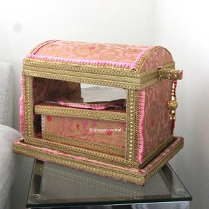 Puede incluir: Una caja decorativa rosa y dorada con una ventana de plástico transparente. La caja está decorada con ribetes dorados y tela rosa. La caja está sentada sobre una mesa de vidrio. El texto "@shagancentral" es visible en la caja.