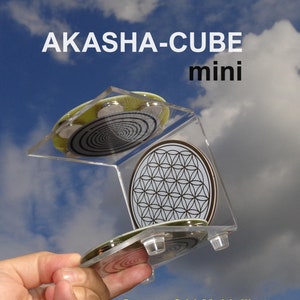 Może przedstawiać: Przezroczysty stojak akrylowy trzyma dwa okrągłe, srebrne i czarne dyski z geometrycznymi wzorami. Górny dysk ma wzór Kwiatu Życia. Dolny dysk ma wzór spiralny. Stojak ma nadrukowany tekst "AKASHA-CUBE mini" i "Quantenfeld-Multiplikator".
