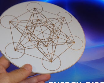 May include: A white round disc with a gold geometric design, featuring a star shape with overlapping circles. The disc is being held in a hand and the text "ENERGY-DISC" is visible below the disc.