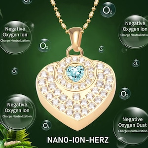 Puede incluir: Un collar con un colgante en forma de corazón de oro con un cristal azul en el centro. El colgante está rodeado de cristales más pequeños y el collar está en una cadena de oro. El texto "NANO-ION-HERZ" está impreso debajo del colgante.