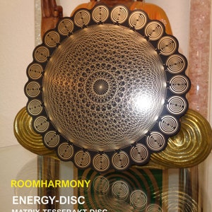 Può includere: Un disco energetico nero e oro con un complesso motivo geometrico, con il testo "ROOMHARMONY ENERGY-DISC" e "240 mm XL Plate". Il disco è esposto davanti a una statua di Buddha in legno. Il diametro è di circa 24 cm.
