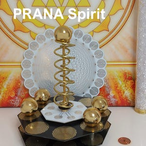 May include: A decorative object featuring a central gold-colored metal coil topped with a sphere, surrounded by four smaller gold spheres on a black hexagonal base. The base displays circular patterns, and the text PRANA Spirit is visible.