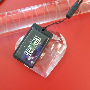 May include: A black rectangular pendant with a spiral design and a black cord. The pendant is on top of a clear heart-shaped crystal. The background is a solid red color.