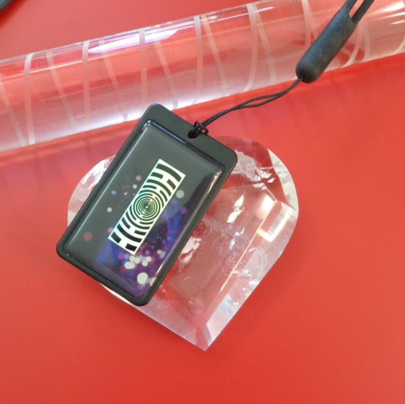 May include: A black rectangular pendant with a spiral design and a black cord. The pendant is on top of a clear heart-shaped crystal. The background is a solid red color.