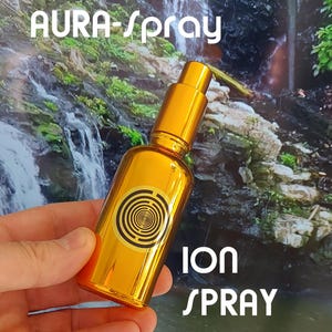 Puede incluir: Un frasco rociador dorado con el texto "AURA-spray" y "ION SPRAY" en blanco. El frasco tiene un diseño circular negro y dorado en la parte delantera. El fondo presenta una cascada y vegetación.