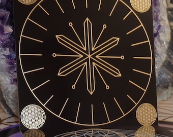 May include: A black square tile with a gold geometric design in the center. The design is a starburst with six points. The tile has the words "WASSER-HEXAGONAL" at the top. There are four smaller gold geometric designs in the corners of the tile.