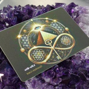 May include: A rectangular card with a detailed design featuring an eye, pyramid, key, and geometric patterns in gold and white on a dark background. The card is resting on a bed of purple amethyst crystals.