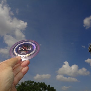 Op de afbeelding: Een ronde, iriserende sticker met Hebreeuwse tekst en een paars-zilveren ontwerp. De sticker wordt in een hand tegen een blauwe lucht met witte wolken gehouden.