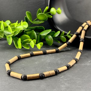 May include: A necklace with light brown wooden beads and black lava rock beads. The necklace is set against a dark backdrop, with a green plant and a black decorative element.