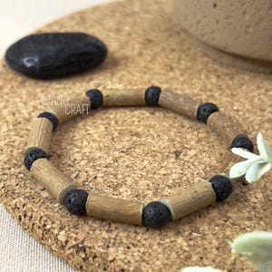 May include: A wooden bracelet with black lava rock beads. The bracelet features cylindrical wooden beads and round black beads. The bracelet is displayed on a cork surface. The text "EVANO CRAFT" is visible.