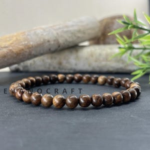 May include: A wooden bead bracelet with dark brown, round beads. The bracelet is displayed on a dark gray surface with a blurred background of light-colored stones and green foliage. The text "EVANOCRAFT" is partially visible.