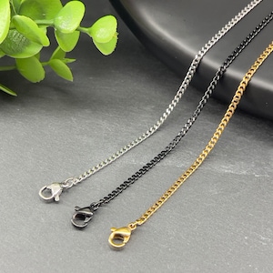 May include: Three curb chain necklaces in silver, black, and gold. Each chain has a lobster clasp closure. The chains are displayed on a dark grey surface with a green plant in the background.