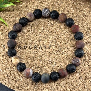 Peut inclure: Un bracelet de perles circulaire composé d'une alternance de perles de lave noire poreuses et de perles de pierre lisses aux tons terreux, dans des nuances de brun, de gris et de beige. Le mot EVANOCRAFT est imprimé en lettres noires au centre.
