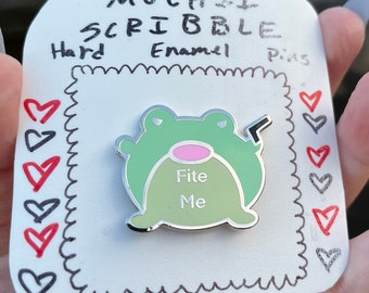 May include: A hard enamel pin featuring a green frog with a pink belly and the words "Fite Me". The pin is on a white card with the words "MOCHII SCRIBBLE Hard Enamel Pins". The card has drawn hearts.