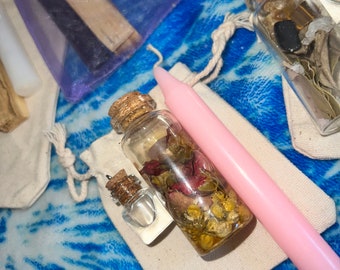 May include: Two small glass bottles with cork stoppers filled with dried flowers and herbs. One bottle is filled with dried rose petals and the other with dried chamomile flowers. A pink candle is also in the image.