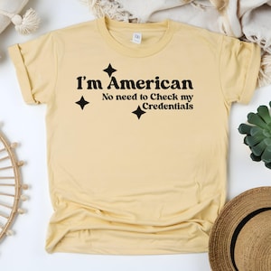 May include: A light yellow t-shirt with the text "I'm American, No need to Check my Credentials" in black. The text is accompanied by three black starburst graphics. The shirt is folded and has rolled-up sleeves.