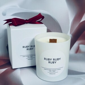May include: A white scented candle with a wooden wick and the text "RUBY RUBY RUBY" on a round label. A matching white box with a burgundy ribbon is next to the candle. The text "HAND POURED SCENTED CANDLE" is also visible.