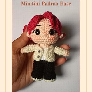 Pode incluir: Uma boneca de crochê com cabelo rosa brilhante, olhos pretos grandes e bochechas rosadas. A boneca veste uma jaqueta creme com dois botões pequenos sobre uma camisa branca e calças pretas. O texto Minitini Padrão Base é visível no topo.
