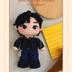 Pode incluir: Uma boneca de crochê com cabelo preto, bochechas rosadas e olhos escuros. A boneca veste uma jaqueta preta, calças azul-escuras e botas pretas. O texto diz: Minitini Stage Persona • Minmin • PDF. A boneca tem um pequeno acessório de borla.