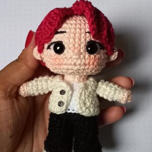 Pode incluir: Uma pequena boneca feita à mão em crochê com cabelo vermelho vivo, rosto cor de pêssego pálido com olhos pretos grandes e bochechas rosadas. A figura veste uma jaqueta branca texturizada com dois botões pequenos sobre uma camisa branca e calças pretas.