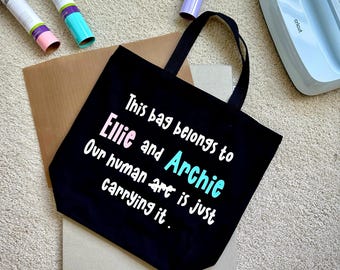 May include: Black tote bag with the text "This bag belongs to Ellie and Archie. Our human art is just carrying it." The bag has black handles and is on a light-colored surface. Craft supplies are visible in the background.