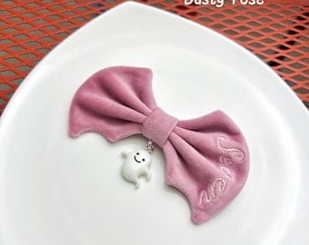 May include: A dusty rose velvet hair bow with bat-wing shaped edges. The bow features a white ghost charm with a smiling face hanging from the center and the word Seven embroidered in a matching dusty rose thread on the right side of the bow.